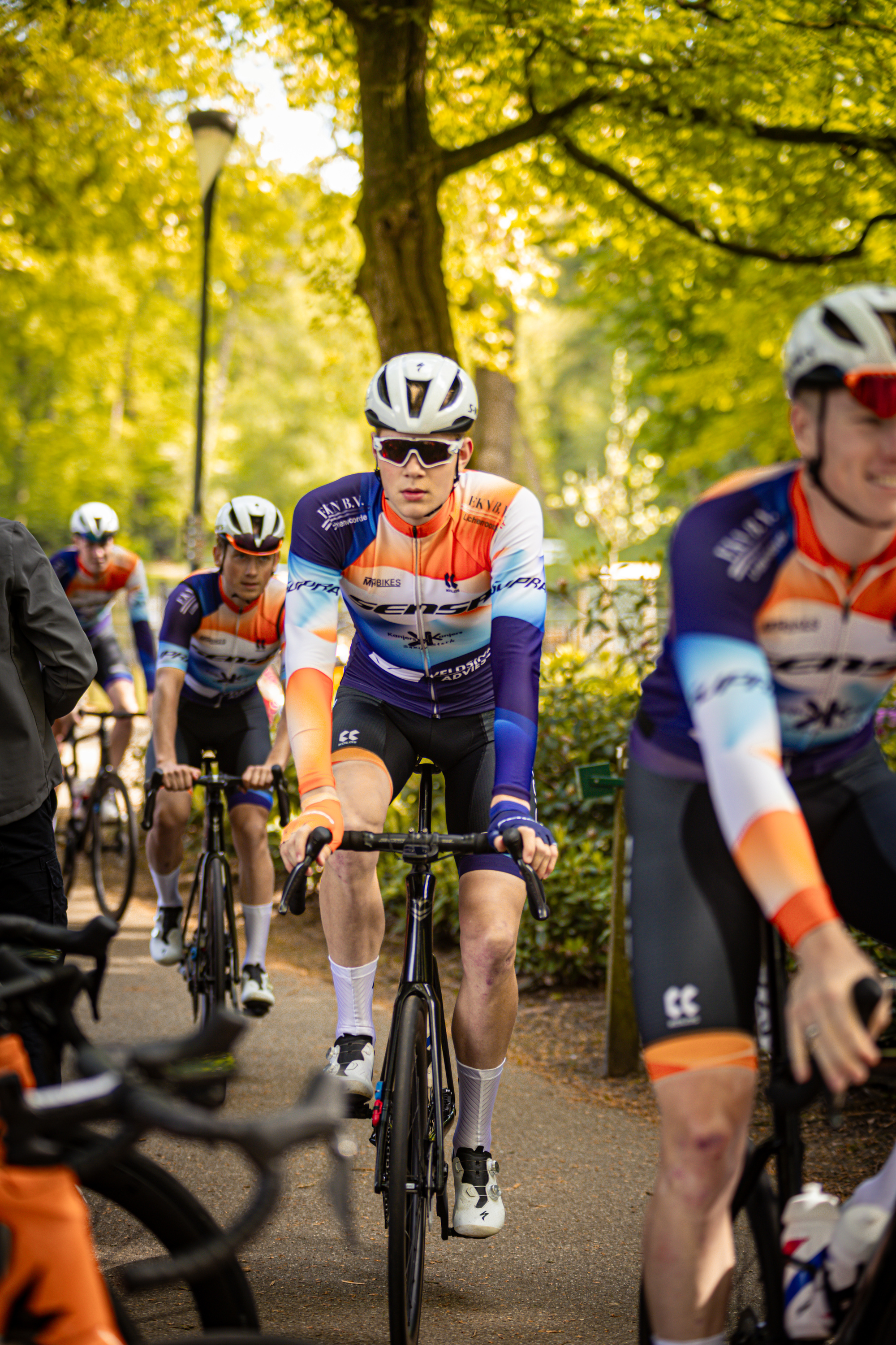 A group of cyclists wearing orange and blue shirts race through a park.