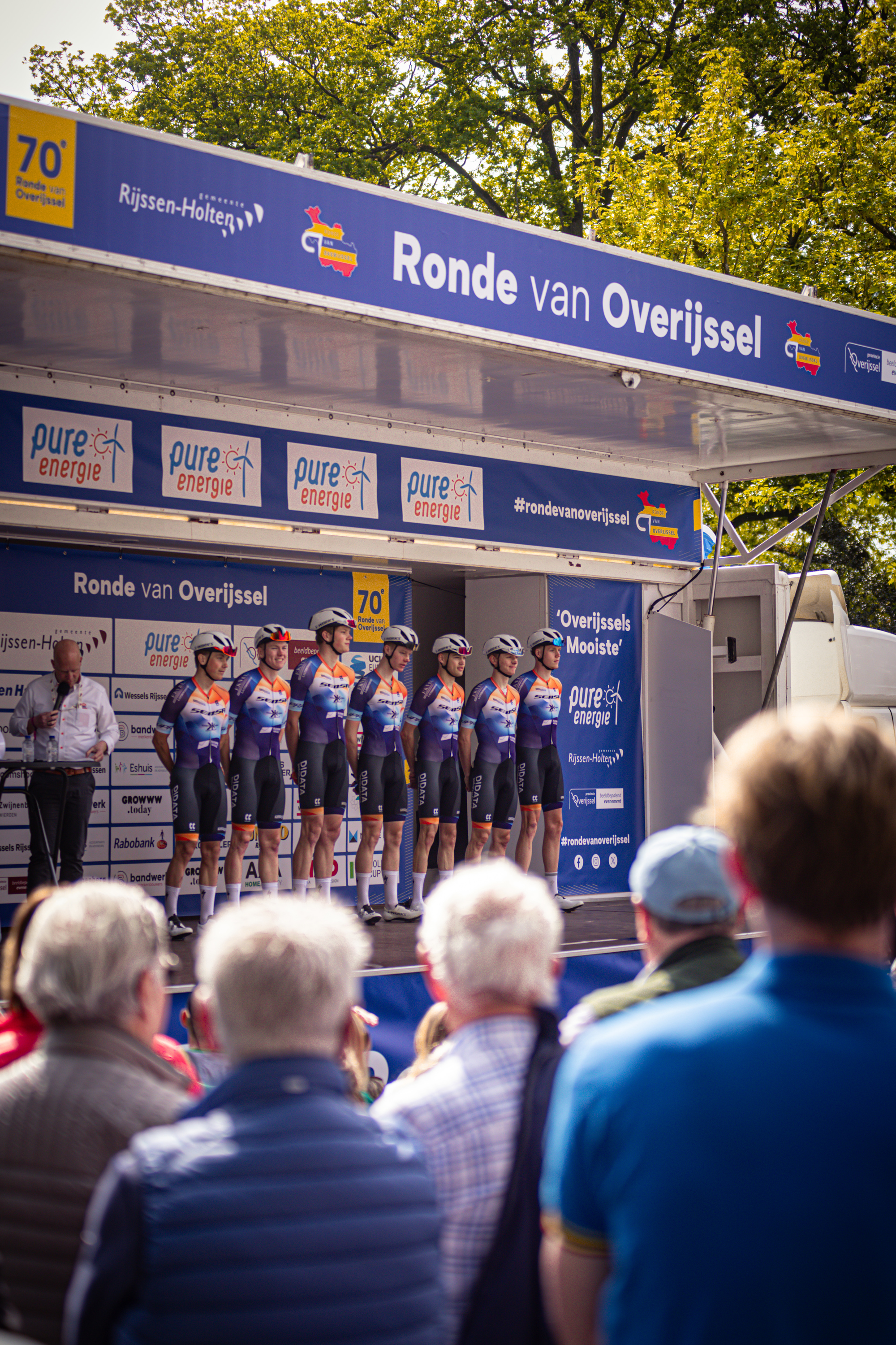 A group of cyclists standing in front of a podium for the Ronde Van Overijssel race, sponsored by Rabobank and Giant.