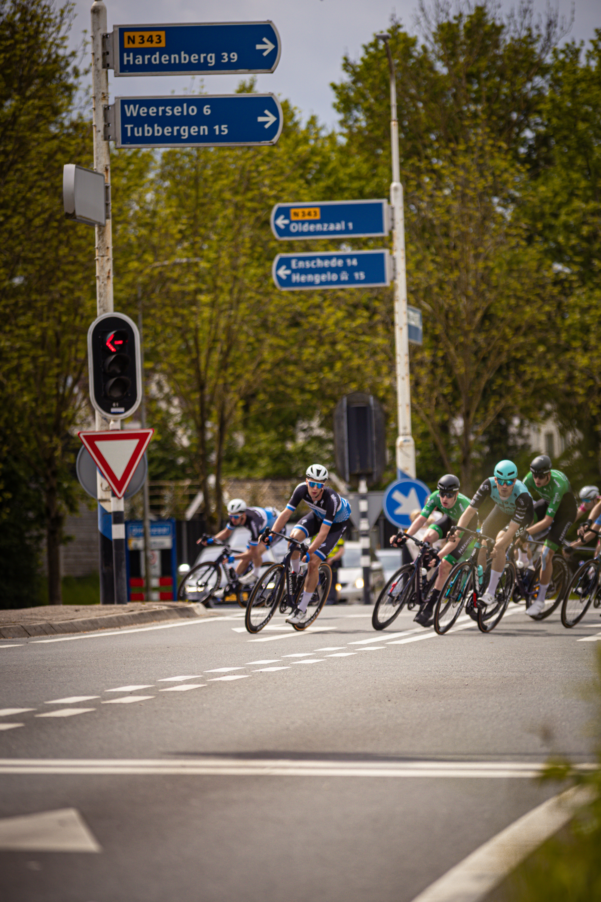 Several cyclists race down a street in the Netherlands, with signs in Dutch above them guiding their way.