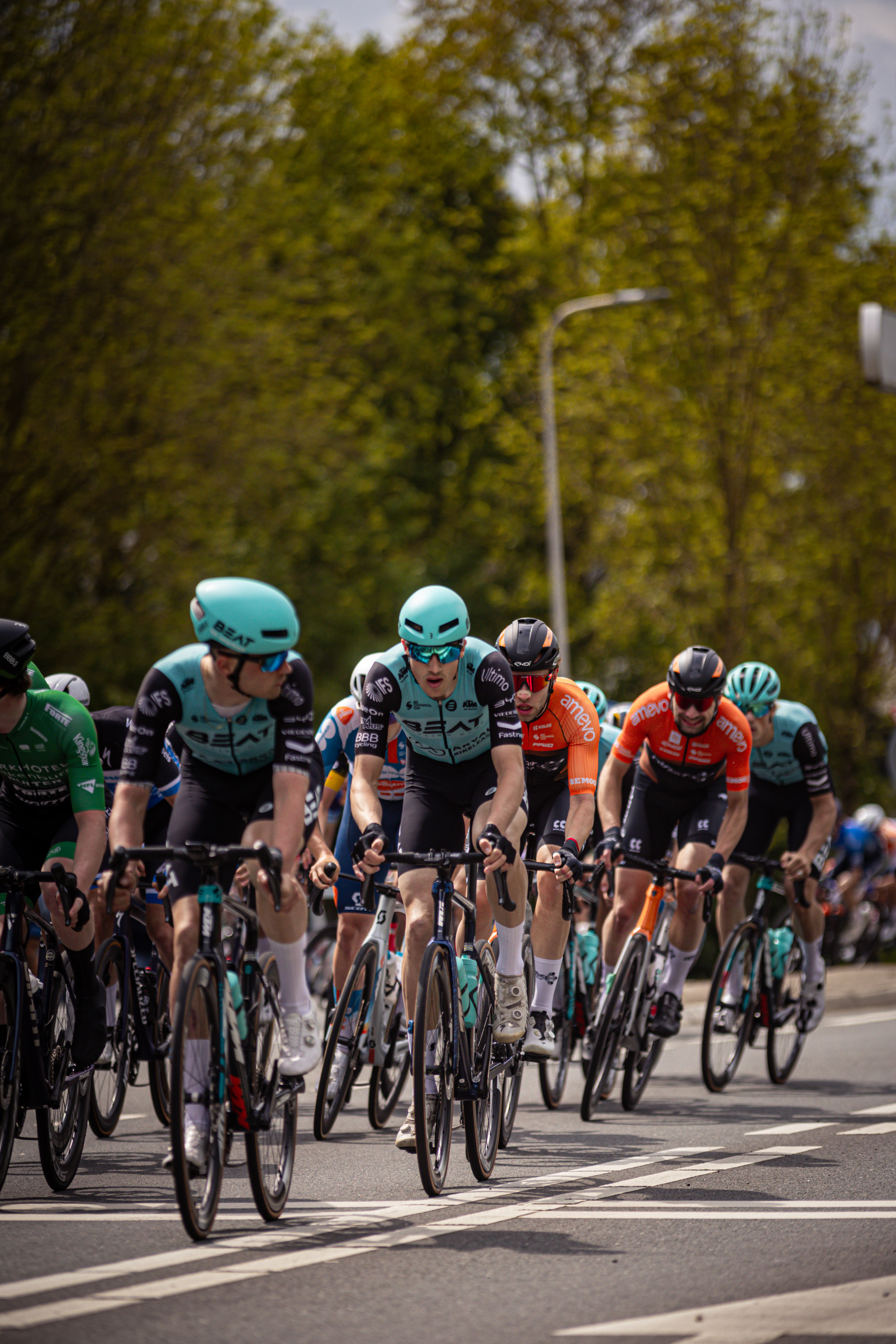 A group of men are riding bikes in a race, one man in the front is wearing an orange jersey with white text.