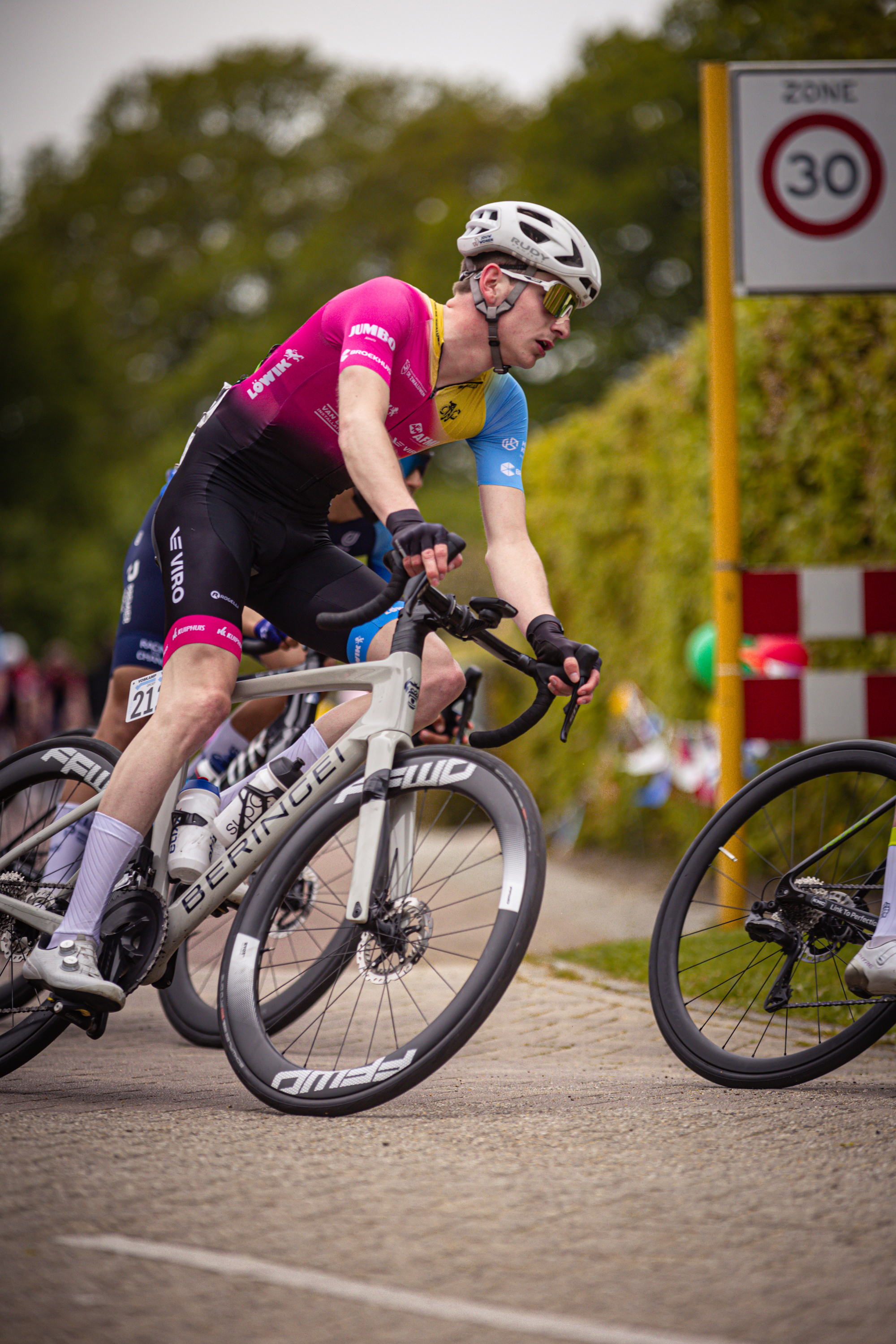 Two cyclists race on a road, one wearing a blue and pink outfit. They are passing two signs with the number 30.