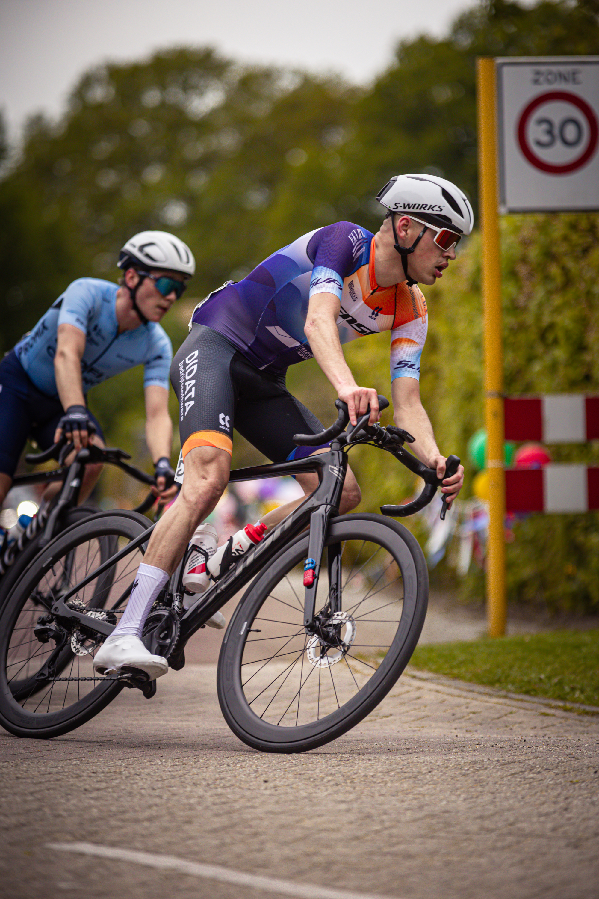 Two cyclists riding down a road with one wearing a Wielrennen jersey.