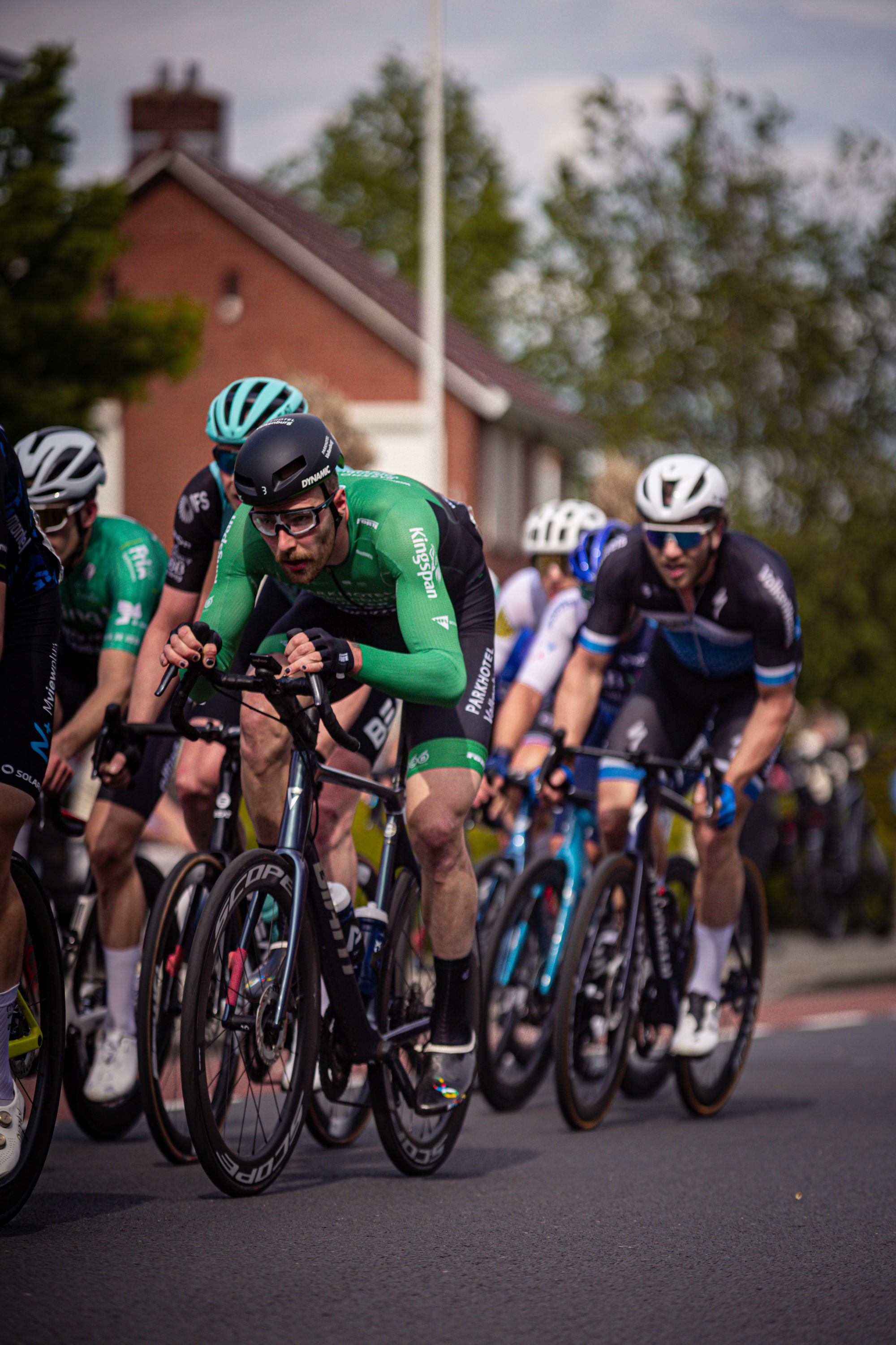 A group of cyclists riding down the street, the one in the lead has a green number 12 on his shirt.