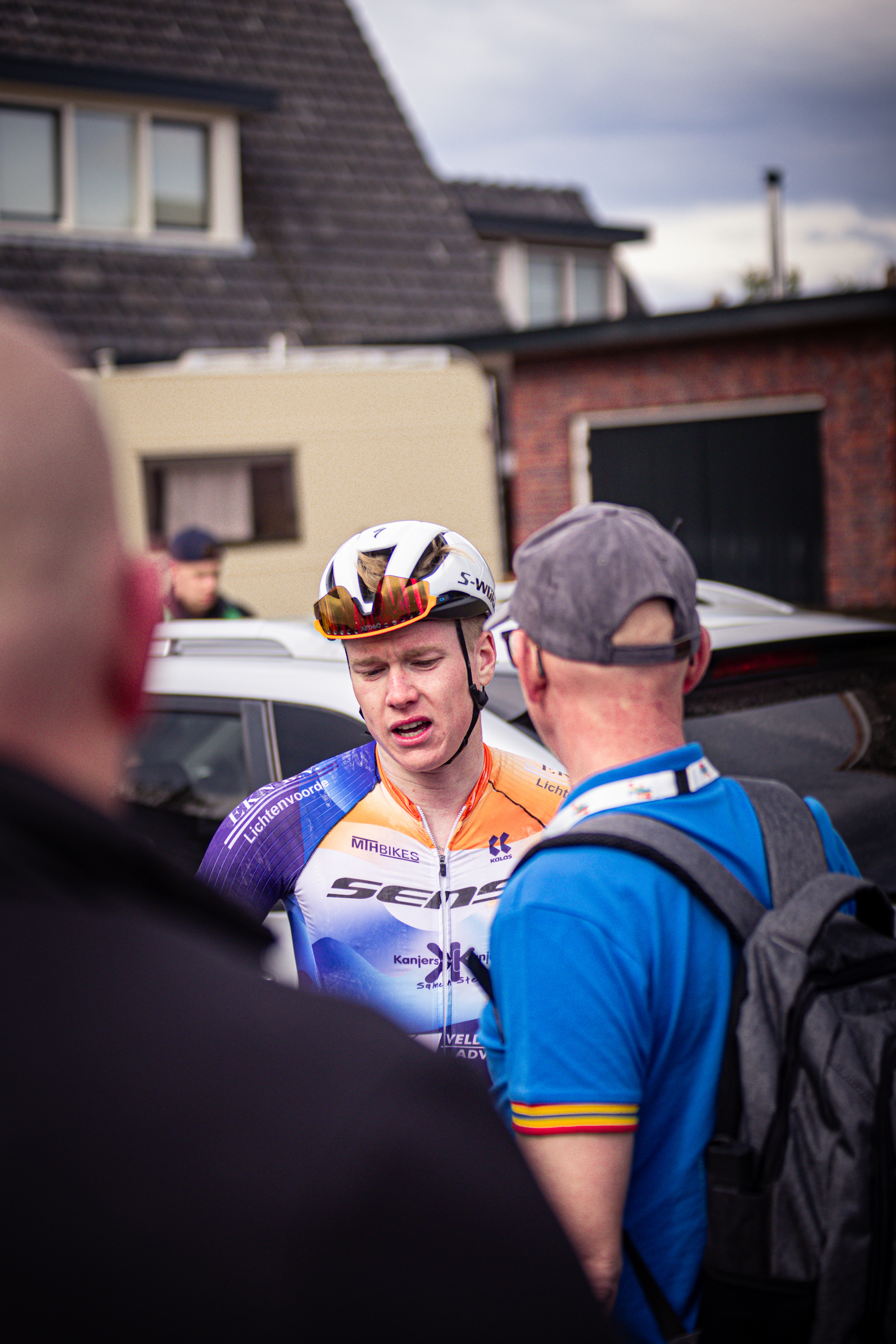 Two cyclists in the street wearing Wielrennen uniforms, one is looking at a man with a blue shirt.