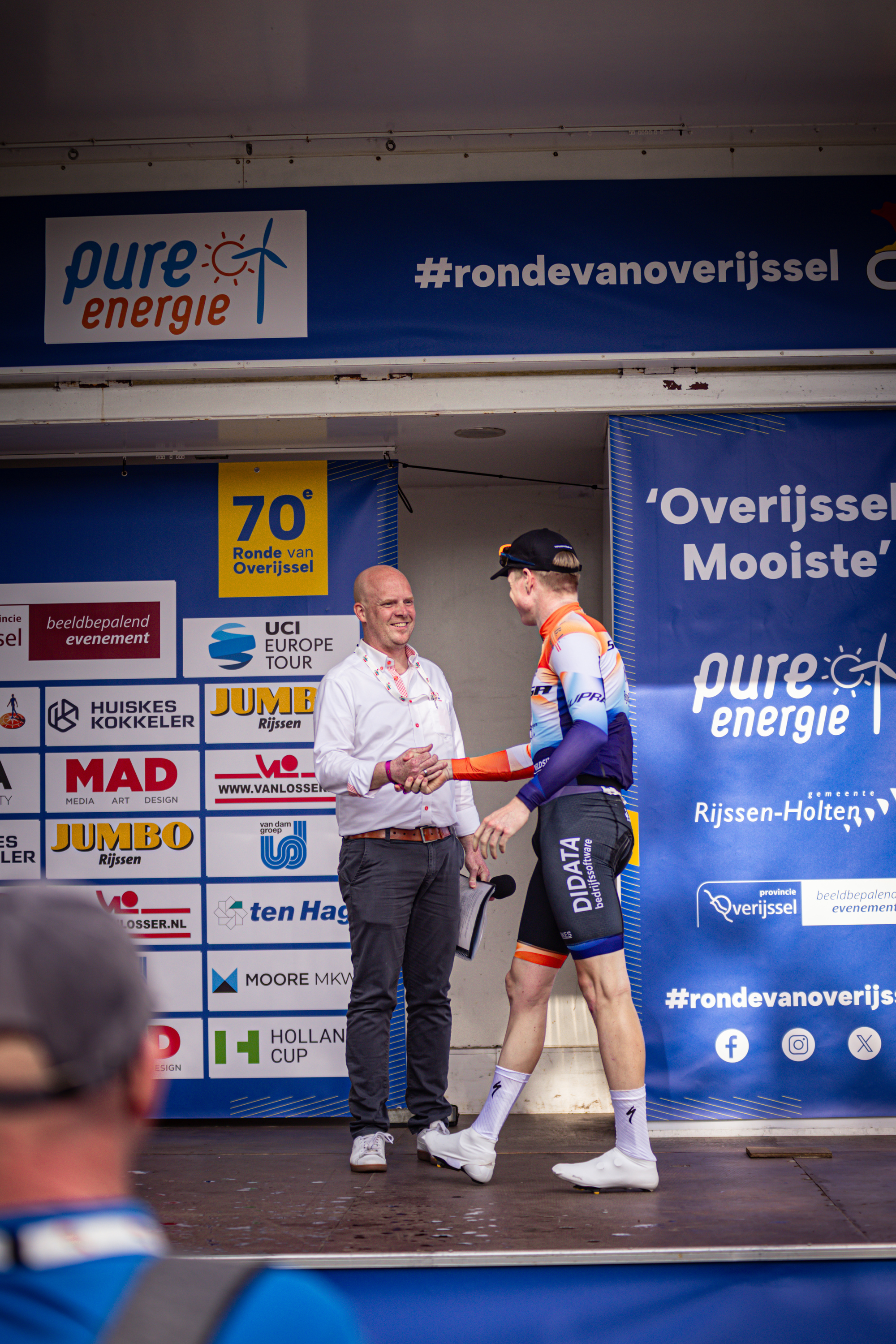 Two athletes shake hands with two men on a blue platform with an advertisement for Rond van Overijssel.