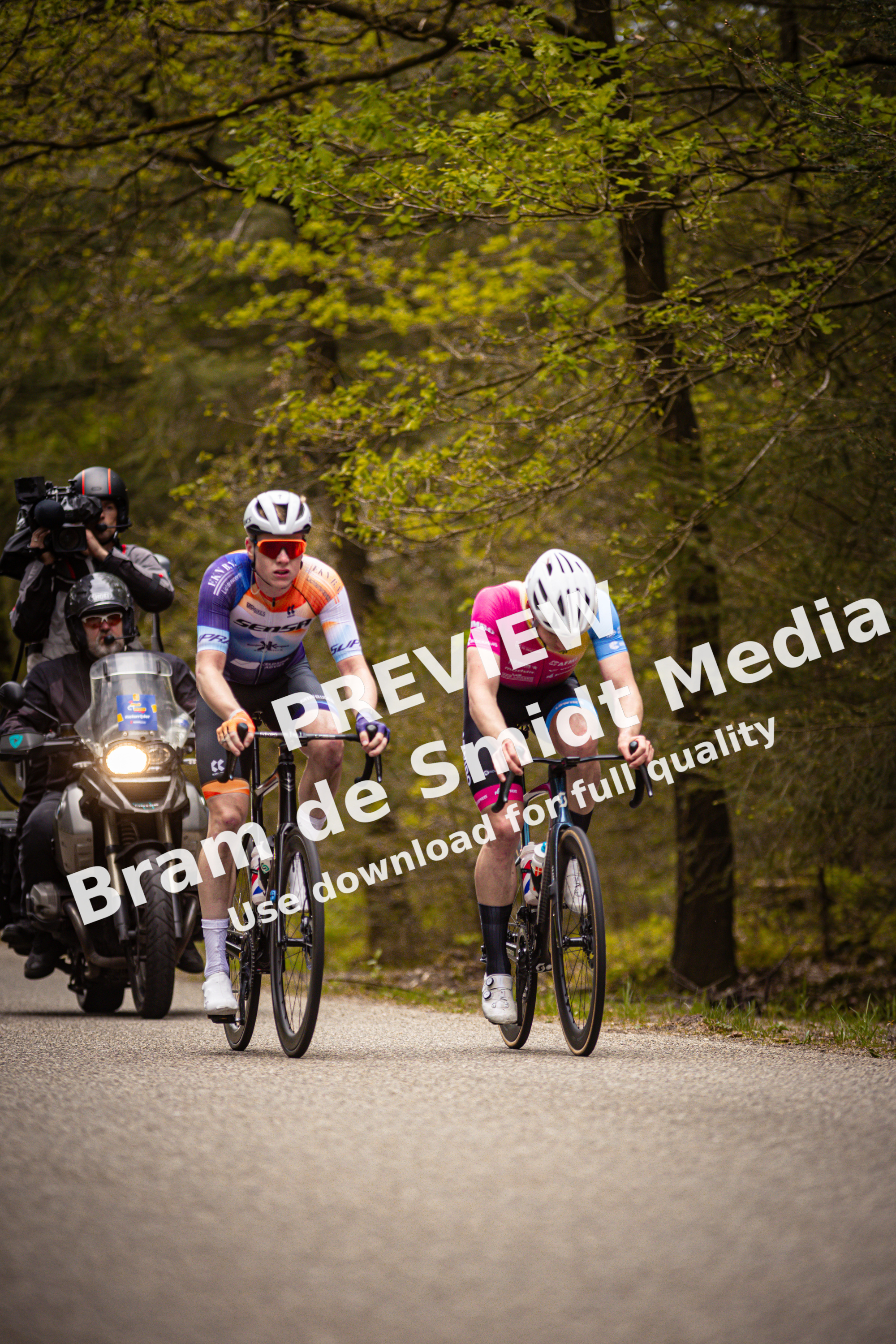 Two men are riding bicycles in a forested area, with one of them wearing a pink jersey.