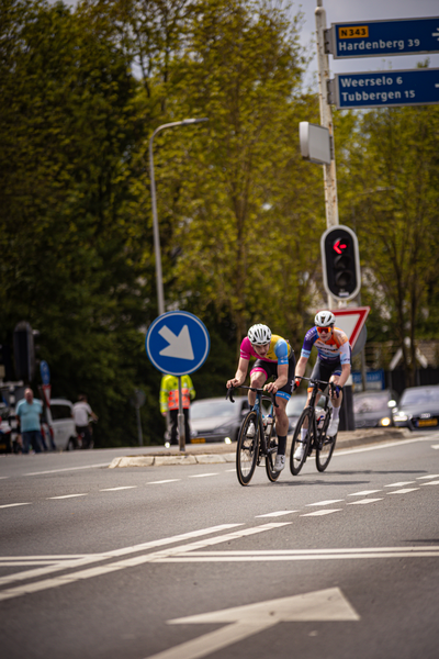 Two cyclists are riding on a street with a sign that says "Wielrennen 2024".