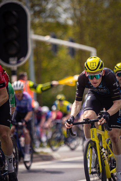 A cyclist with a yellow helmet and blue sunglasses is riding down the road in a bike race.