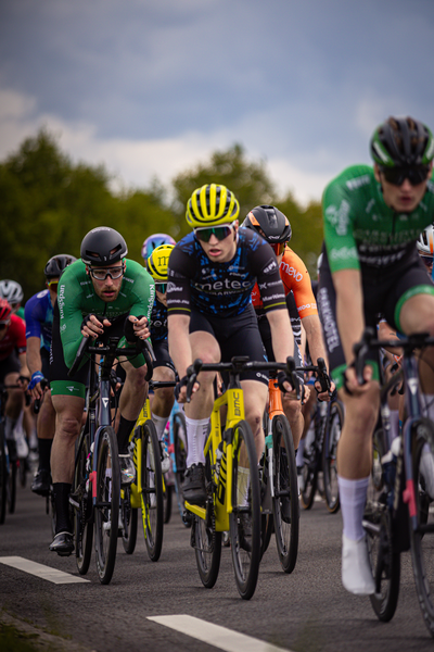 Riders on bicycles are racing in a group with one man wearing a yellow helmet and another wearing a black helmet.