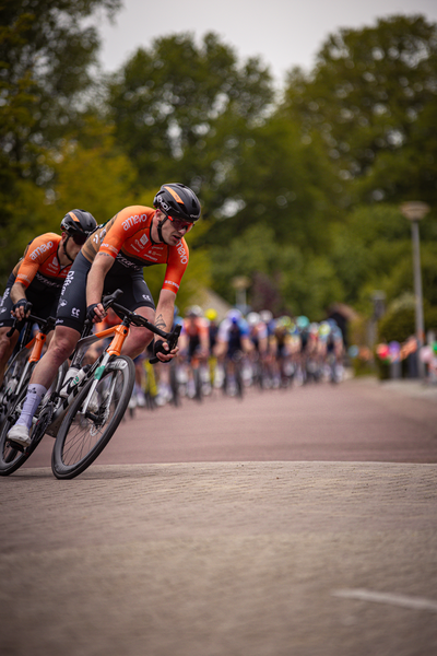 A group of cyclists wearing orange and black jerseys race down a street.