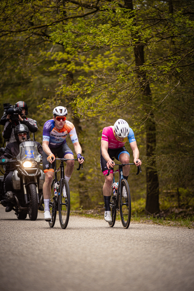 Two men are riding bicycles in a forested area, with one of them wearing a pink jersey.