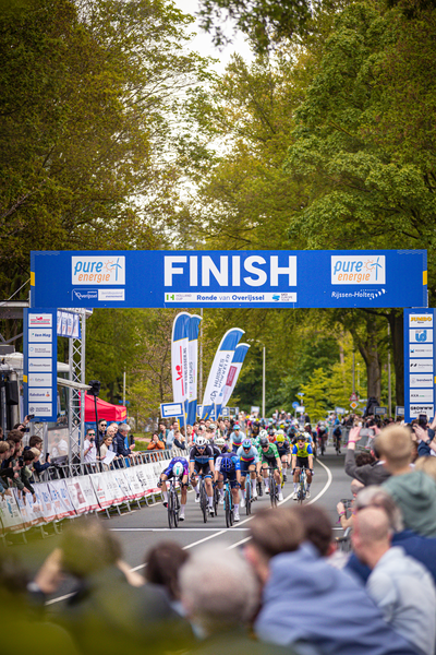 A blue and white banner displays "FINISH RONDE VAN OVERIJSSEL" in front of a crowd of people watching cyclists race.