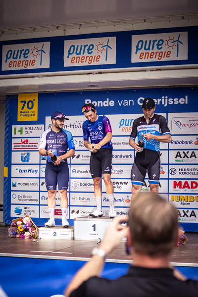 Four men standing at a podium with one man wearing number 1. They are wearing racing outfits and each has a blue hat on.