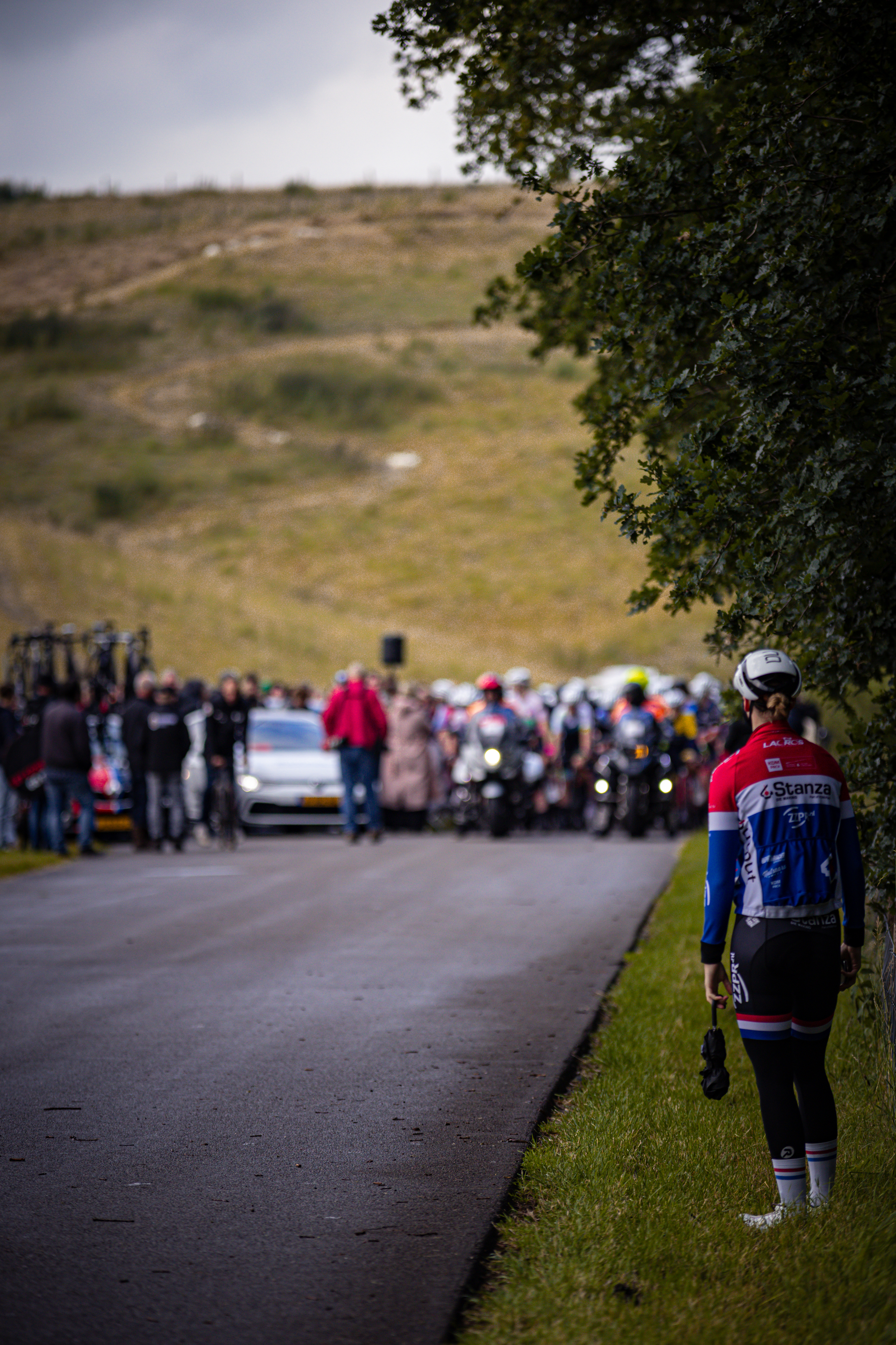 A man in a blue and red jersey that says Dames Elite ZC stands next to the road.