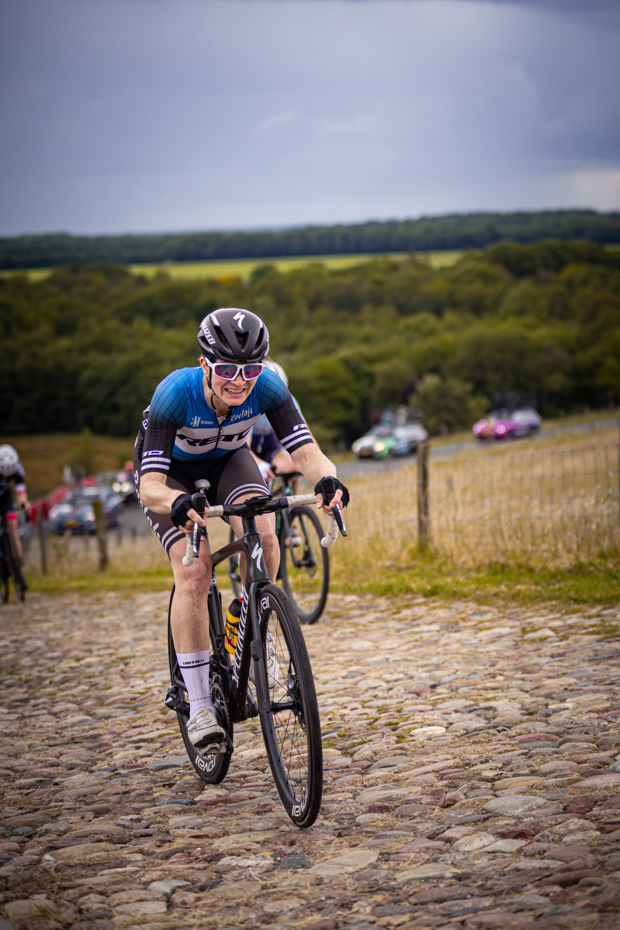 A person in a blue and black jersey wearing sunglasses riding a bike.