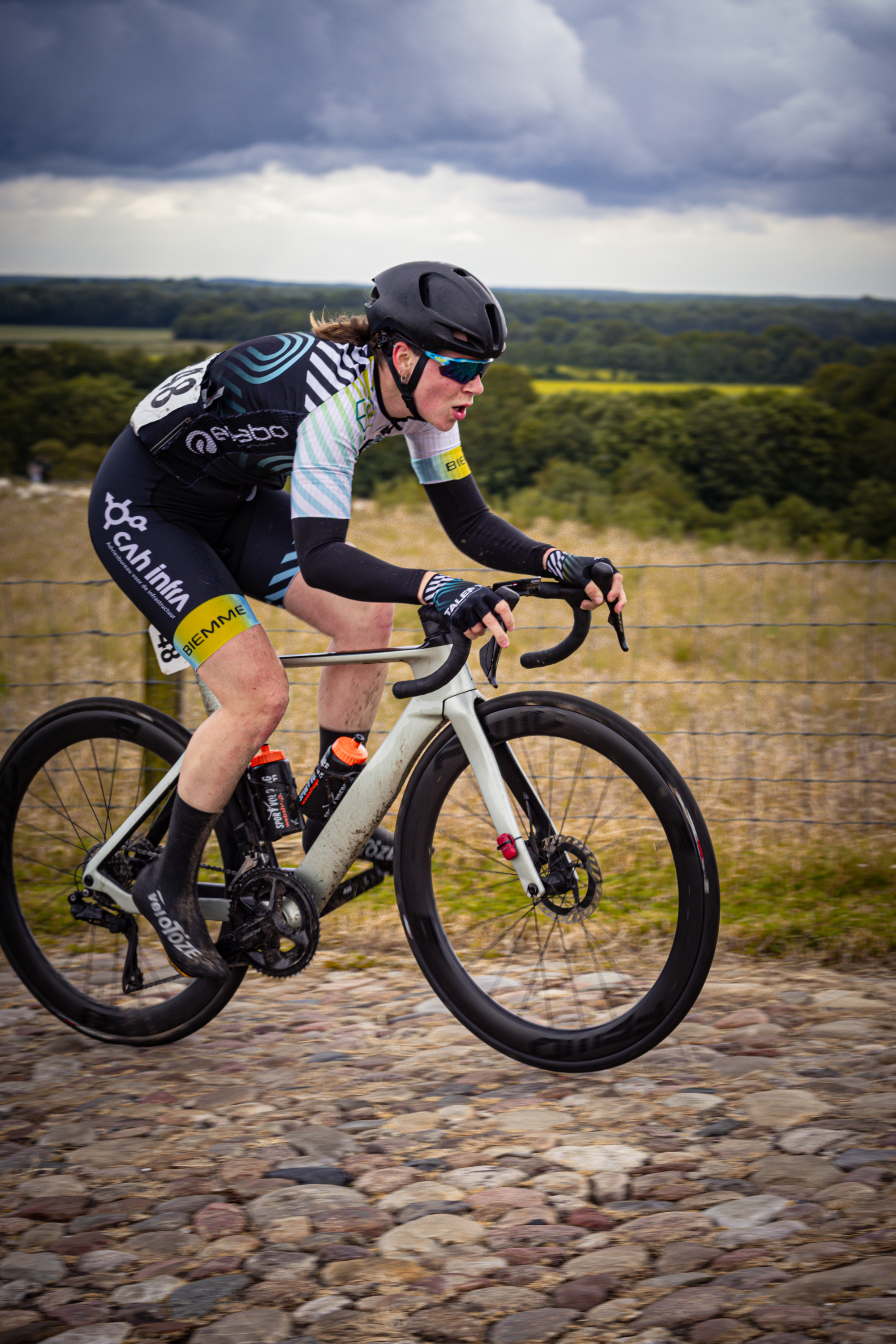 A woman in a jersey and helmet is riding a bike on a cobblestone road.