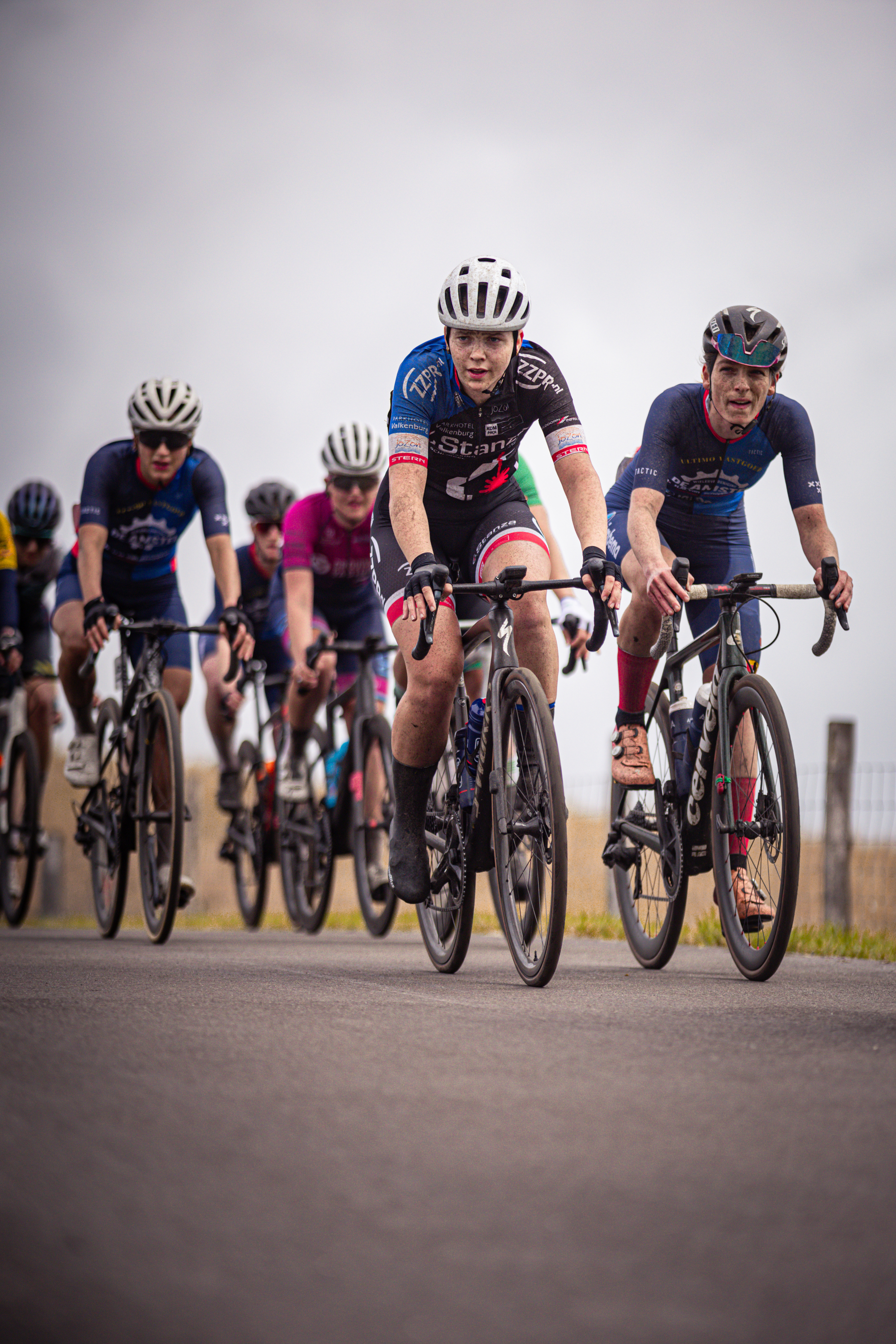 A group of cyclists race down a road, with the cyclist in the lead wearing a black jersey with red and white stripes.