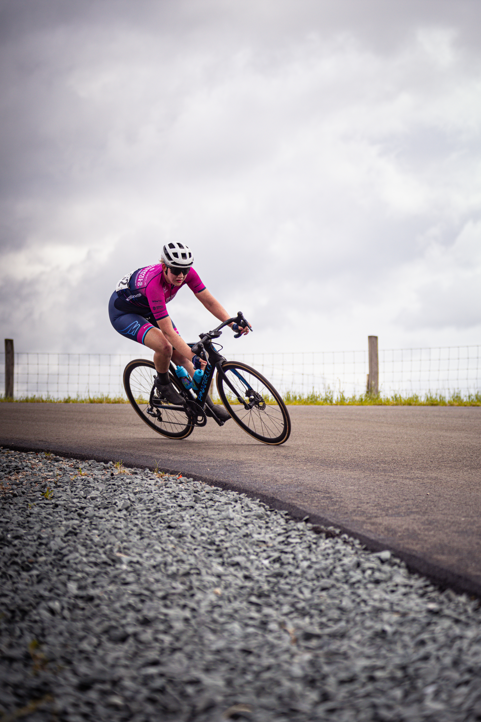 A woman wearing a purple jersey and black helmet rides a bicycle on the road, with a fence visible in the background.