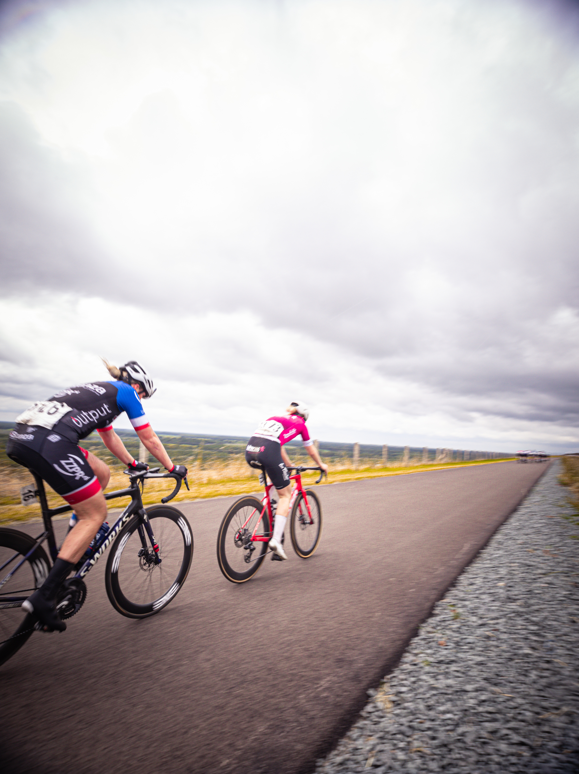 Two cyclists race down a gravel road under a cloudy sky.