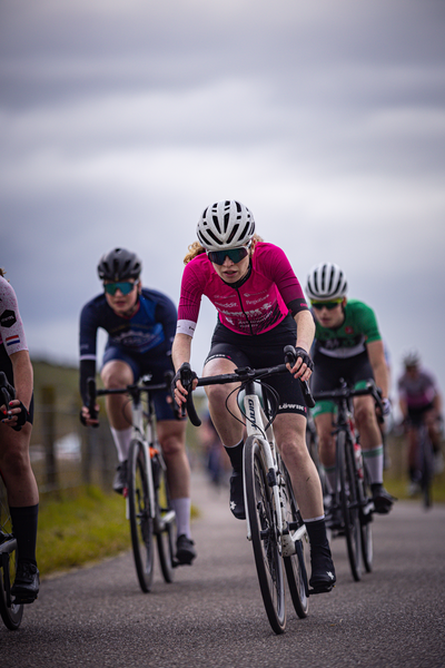 A race of three cyclists in 2024, the woman leading wears a pink jersey with an advertisement for "Hulst".