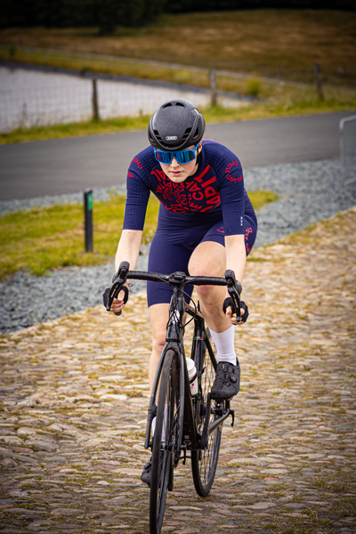 A woman wearing a blue and red cycling outfit rides a black bike on cobblestones.