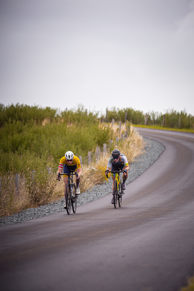 The two cyclists are riding down a road, one of them wearing an "E" on their helmet.