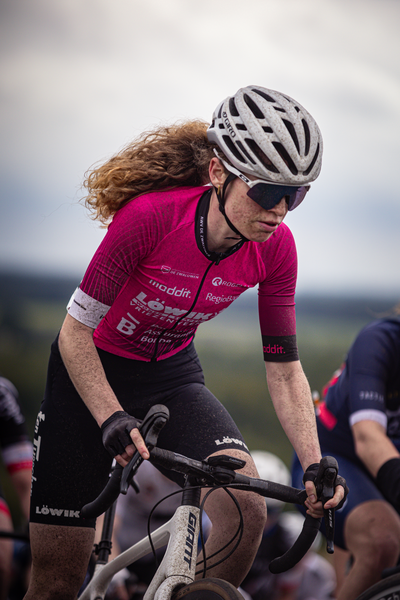 A woman in a red shirt and black pants is riding a bike. She has the words "Nederlands Kampioenschap, 2024" on her chest.