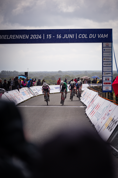 A group of cyclists are riding down a street under an arch with the words Wielrennen 2024 written on it.