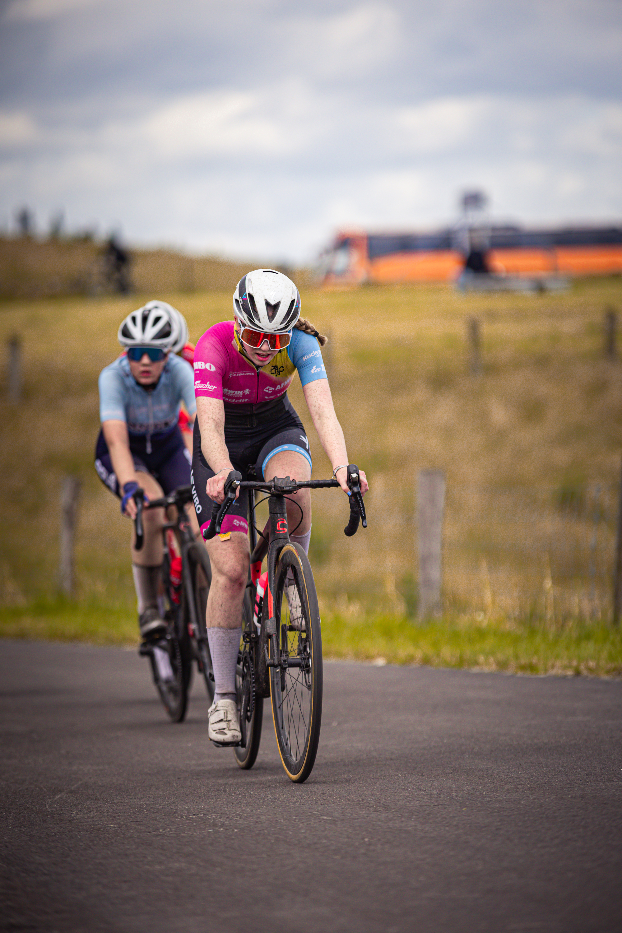 Two women in helmets ride bicycles down a road. The woman in the pink jersey is ahead, leading the race.