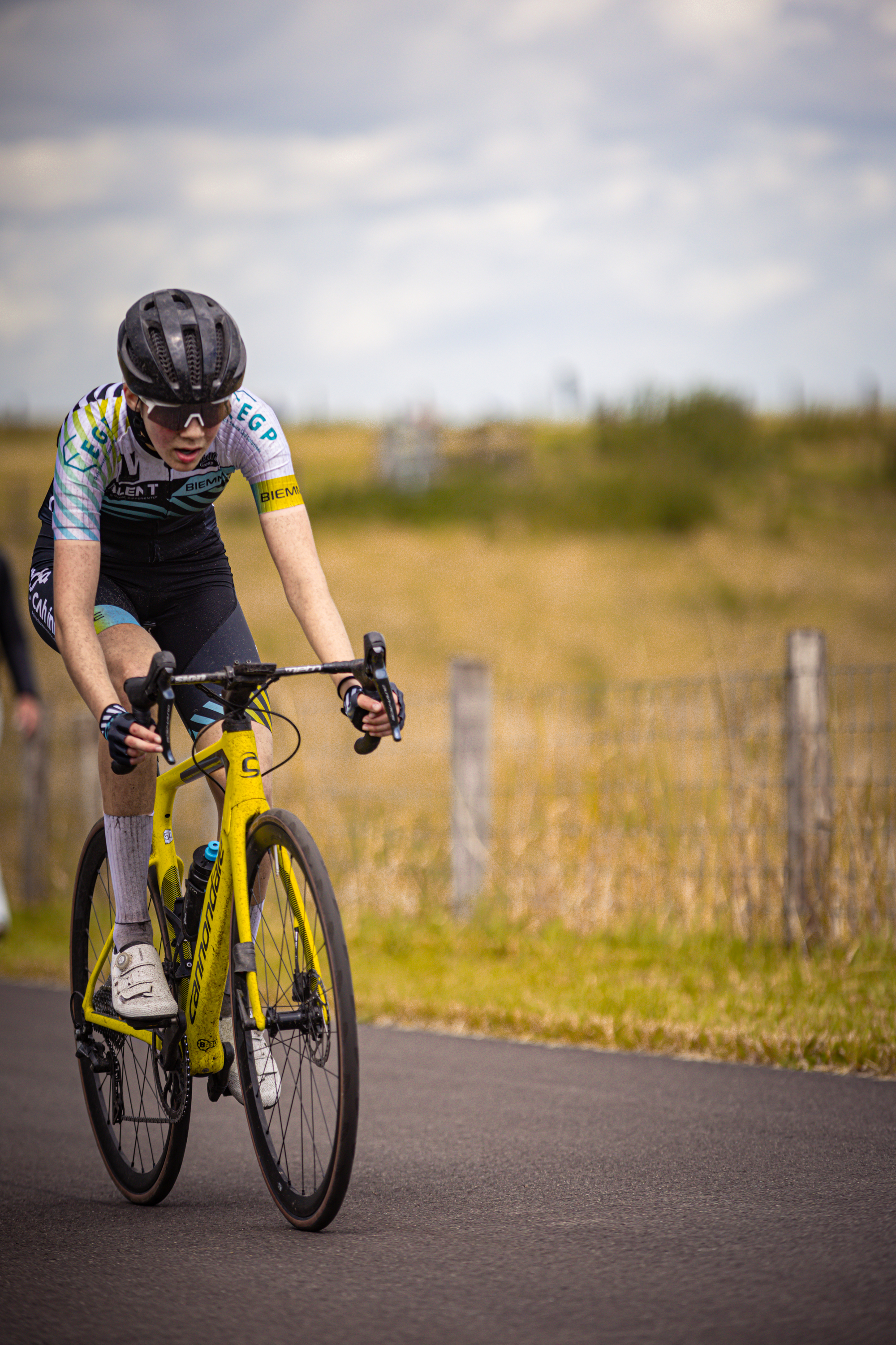 A cyclist wears a blue and white top and helmet. She rides a yellow bike with black handlebars on a road near grass.