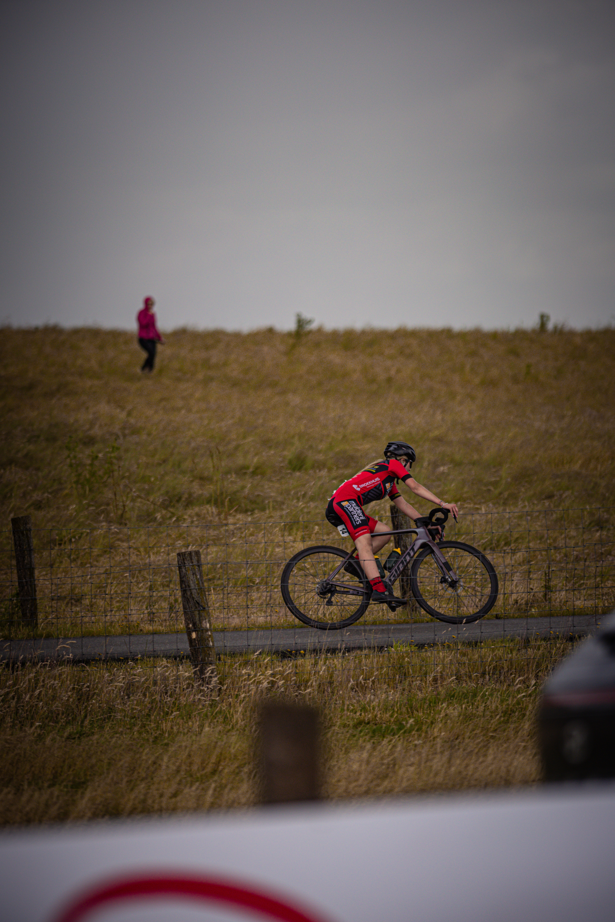 A cyclist wearing red gear is riding down a road near a fence.