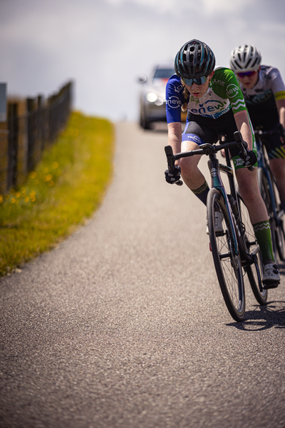 Two cyclists on a road with the person in front wearing a blue jersey that says "Nederlands Kampioenschap".