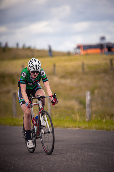 A female athlete wearing a green and black top and white shorts. She is riding her bicycle on a road.