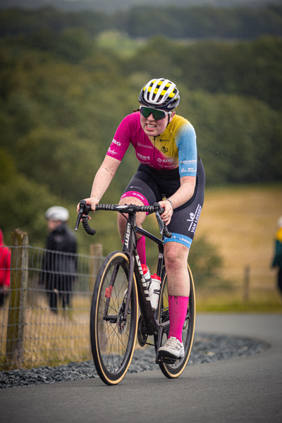 A person wearing a pink and blue jersey, black pants, and a yellow helmet riding a bike down the road.