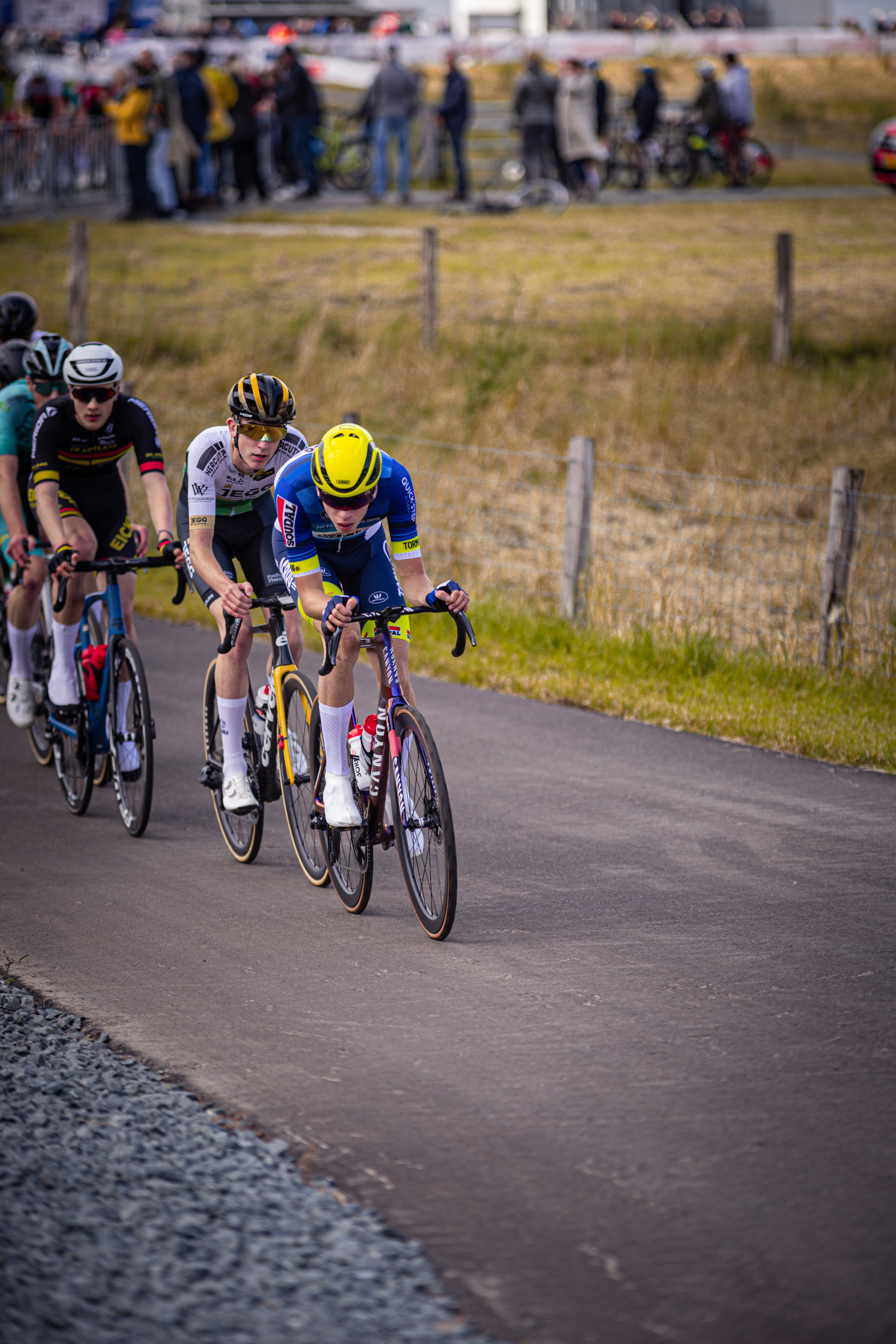 A group of cyclists, wearing blue and yellow helmets, race on a road near the Nederlands Kampioenschap 2024 track.