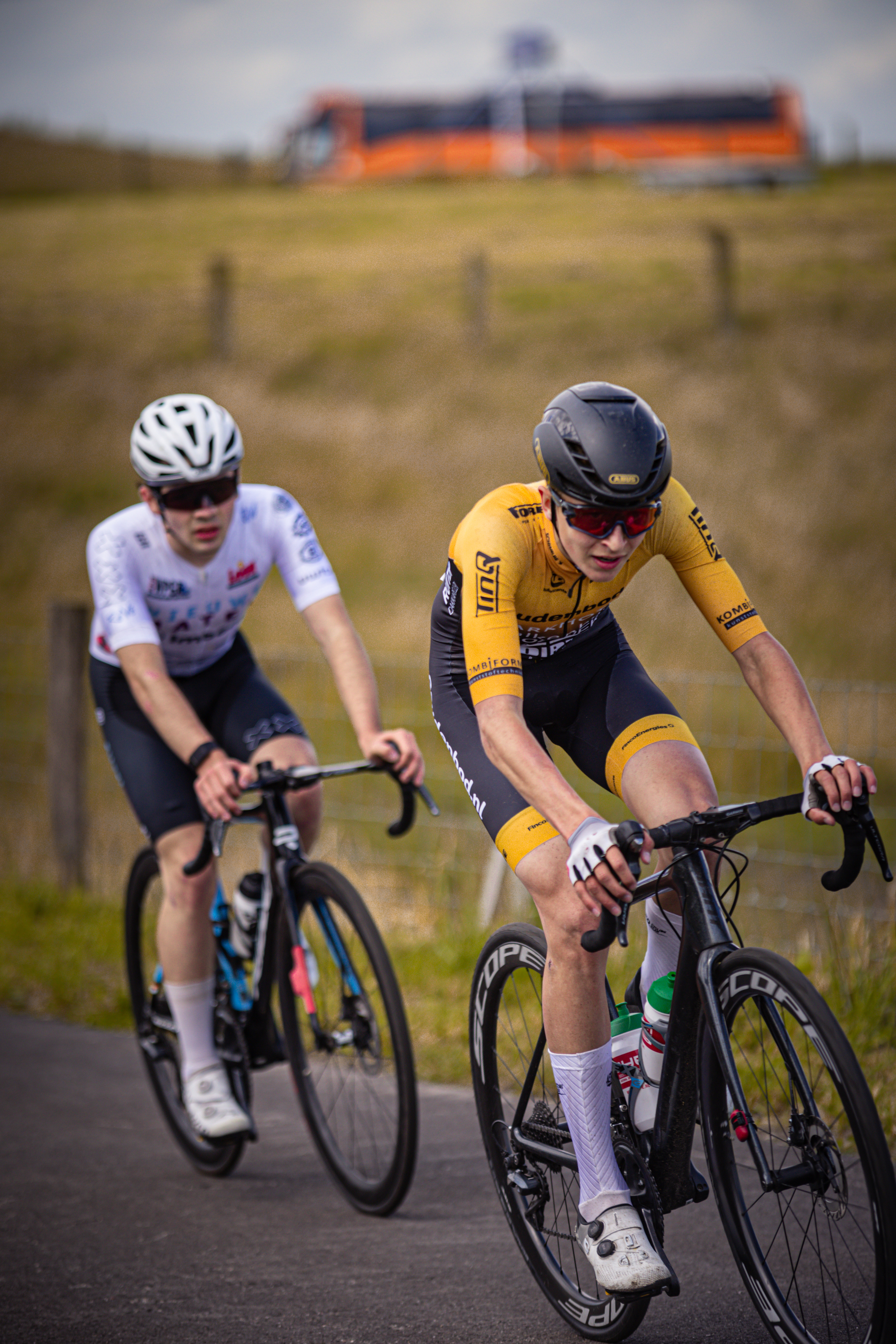 Two cyclists on a road with one of them wearing a yellow shirt that says "Junioren Mannen".