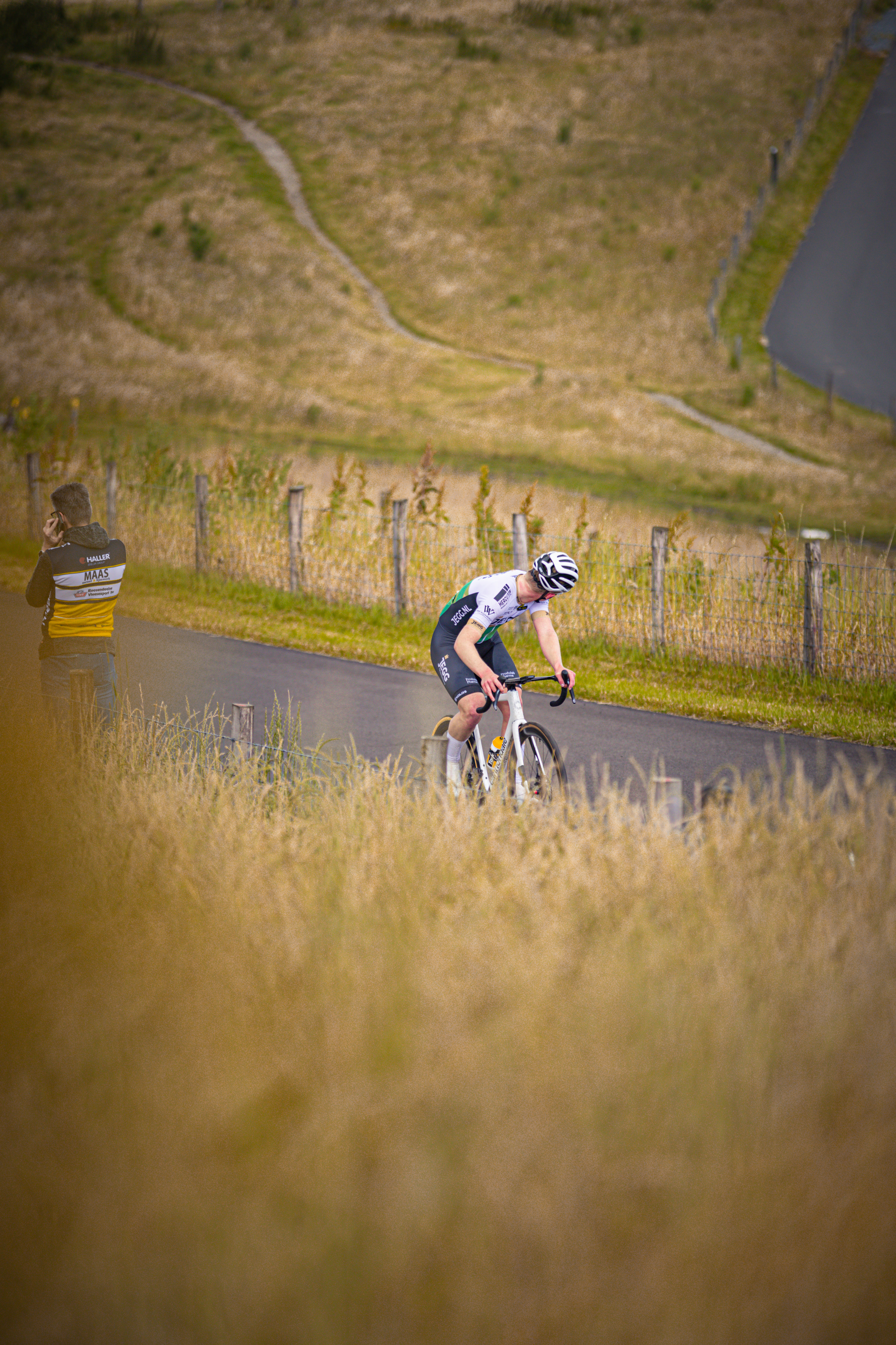 A bicyclist in a black jersey is racing down a dirt road with two other men watching him.