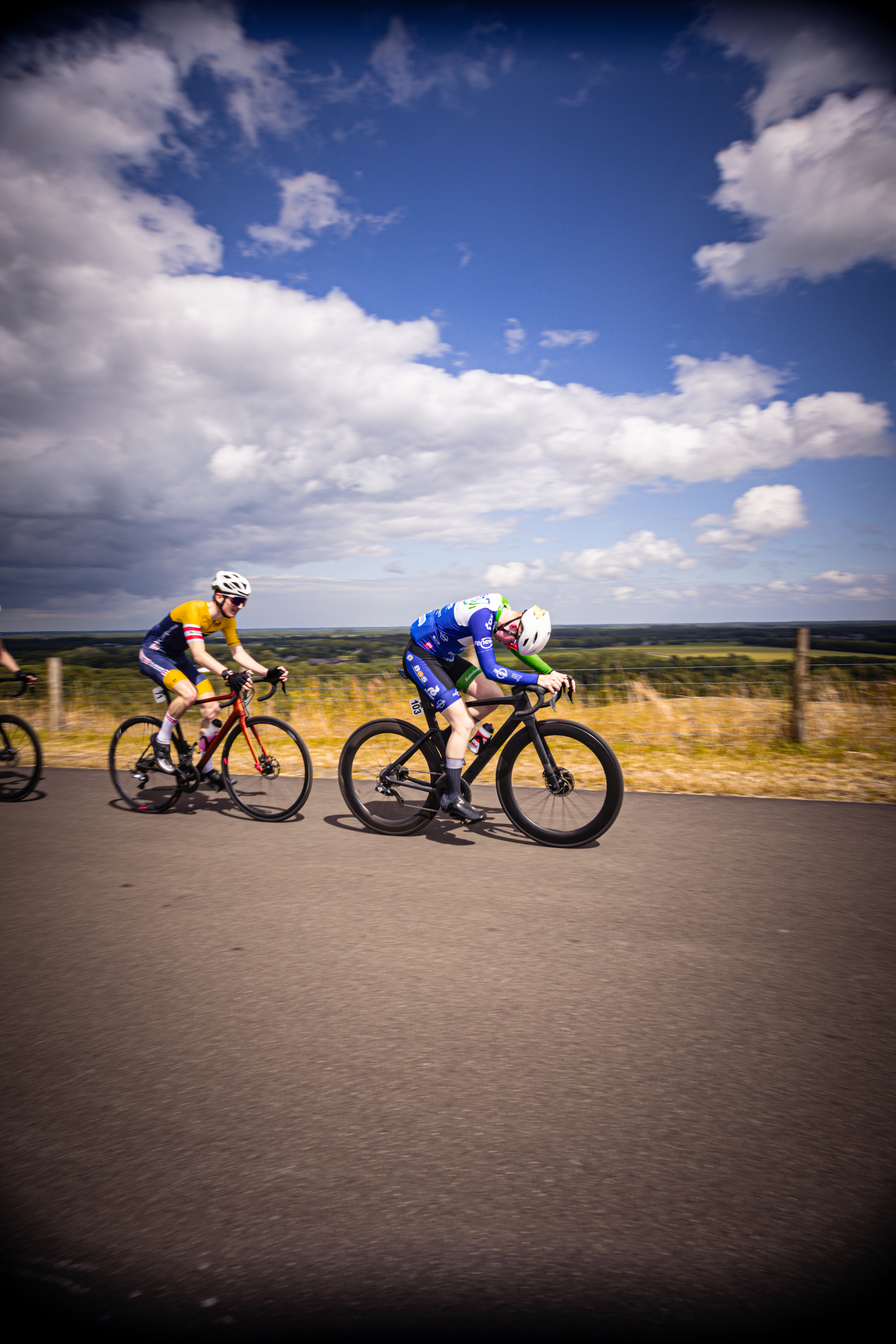 Two cyclists race on a road under blue skies. The cyclist in the lead is wearing a blue and white jersey.