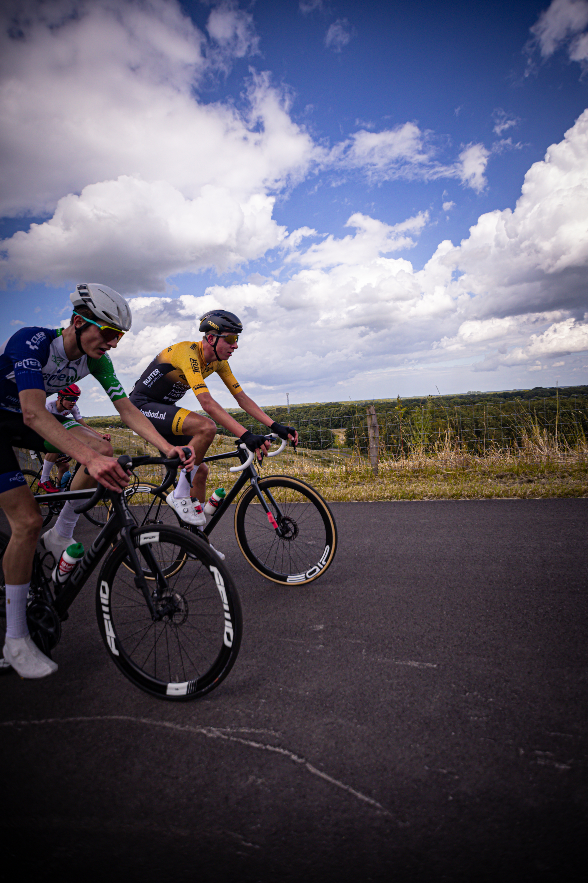 Two men are cycling on a black road with clouds in the sky.