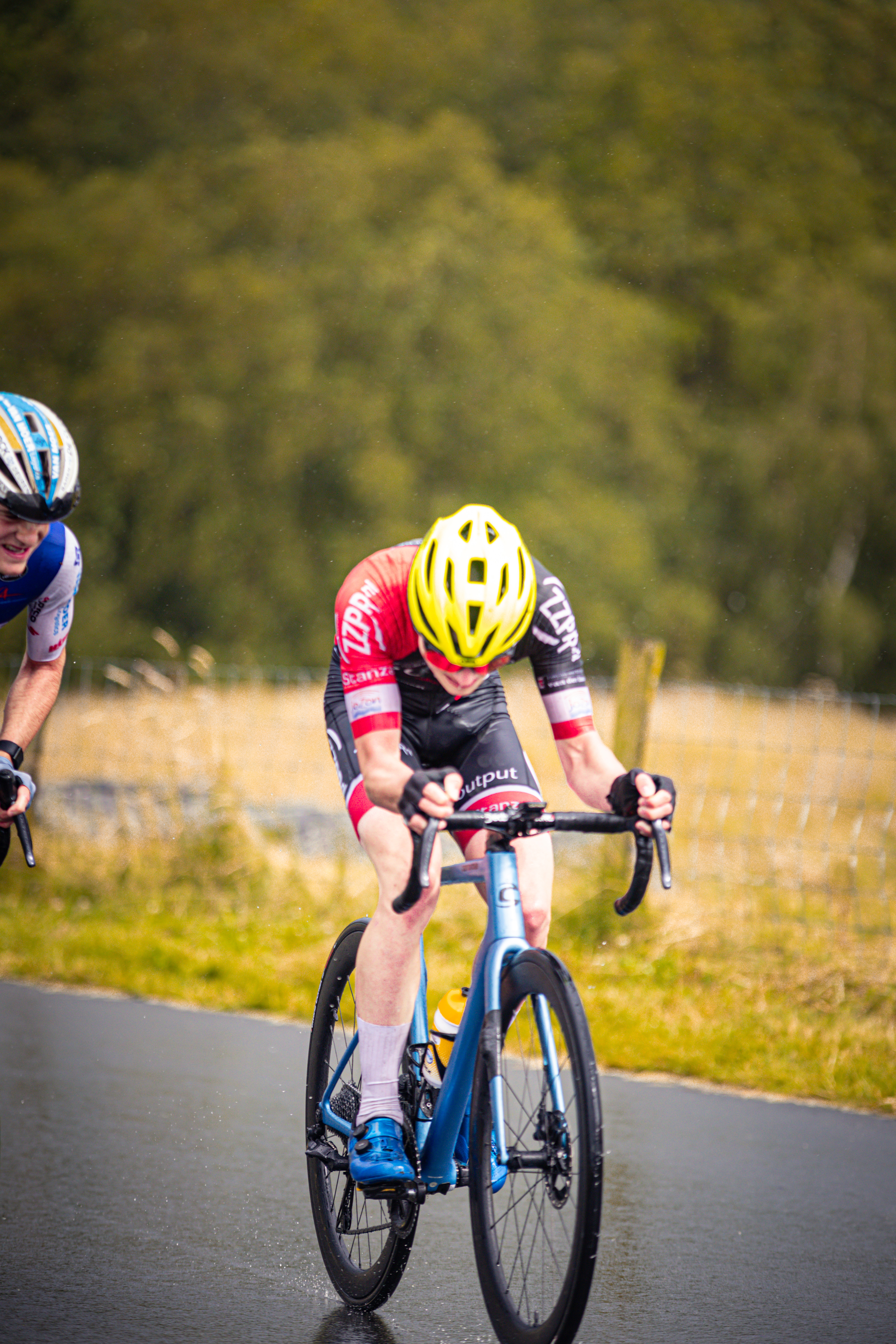 Two cyclists racing on a wet road. One of the cyclists is wearing a yellow helmet with the letter "G" on it.