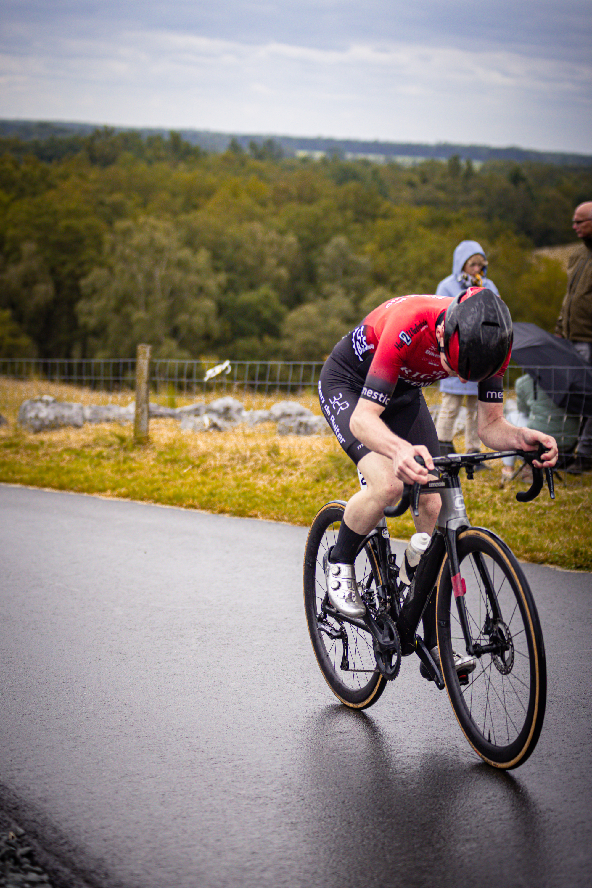 A bike racer with the numbers 8 and 3 on their helmet, wearing a black and red outfit is riding a black bike.