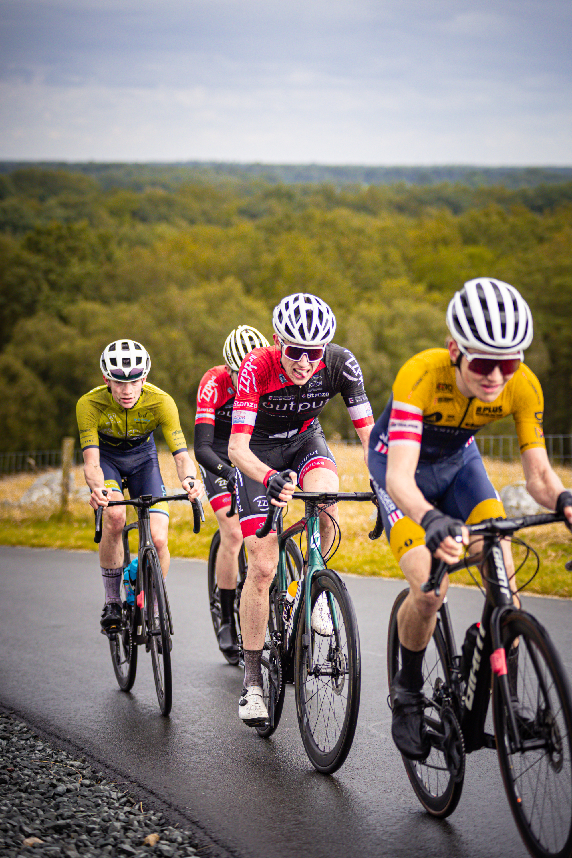 A bike race between three young men is taking place. The man leading the race is wearing a red and black jersey.