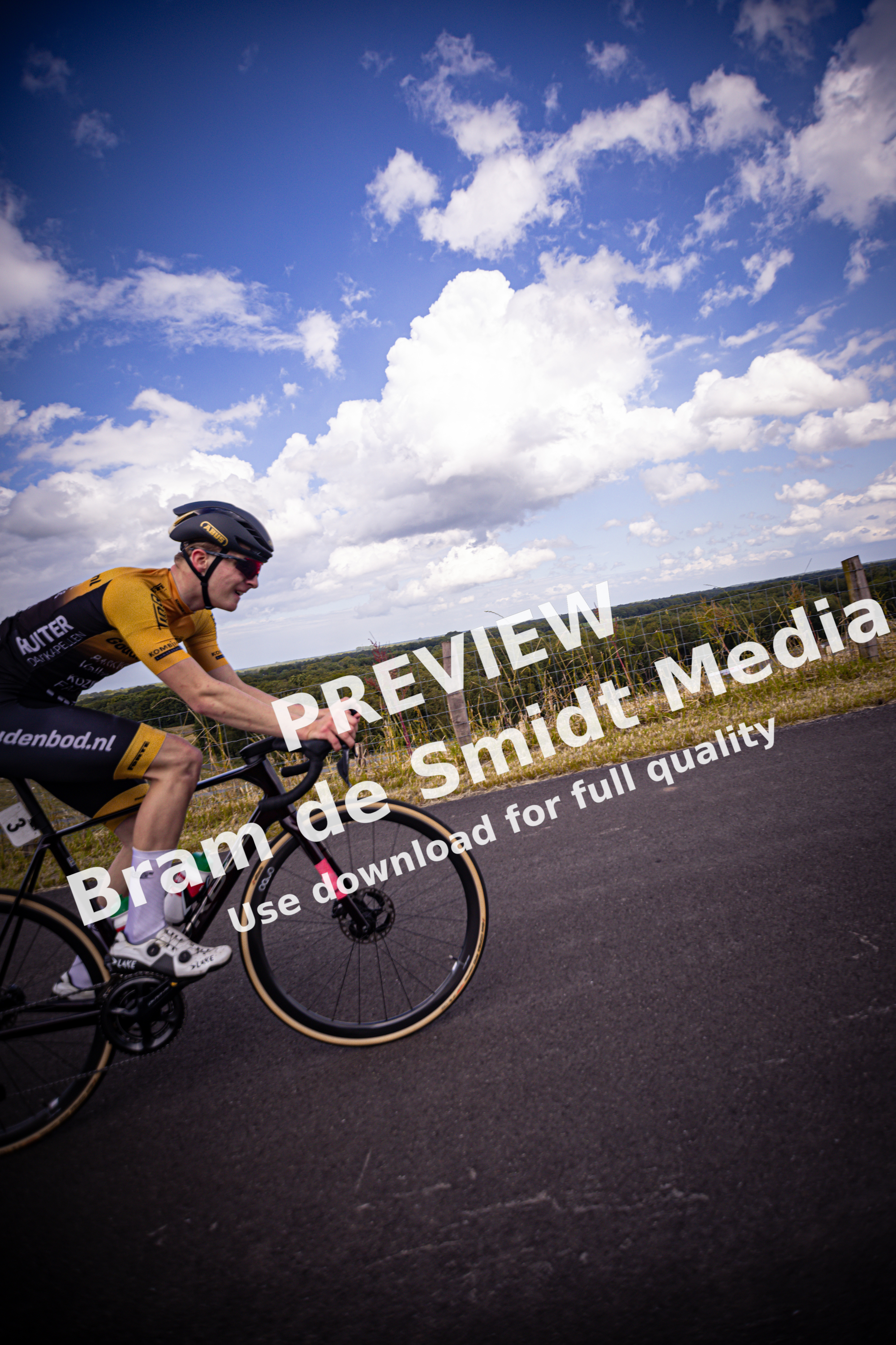 A man in a yellow and black shirt rides a black bike down the road, the word "Nederlands" is visible on his jersey.