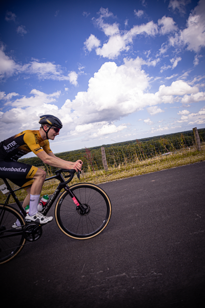 A man in a yellow and black shirt rides a black bike down the road, the word "Nederlands" is visible on his jersey.