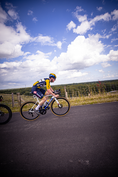 A cyclist in a yellow and blue jersey races down the road, representing Nederlands Kampioenschap.