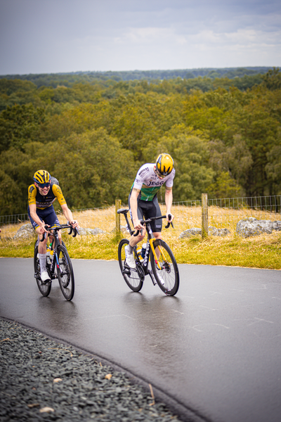 Two cyclists are on a road, one in yellow and black riding to the right.