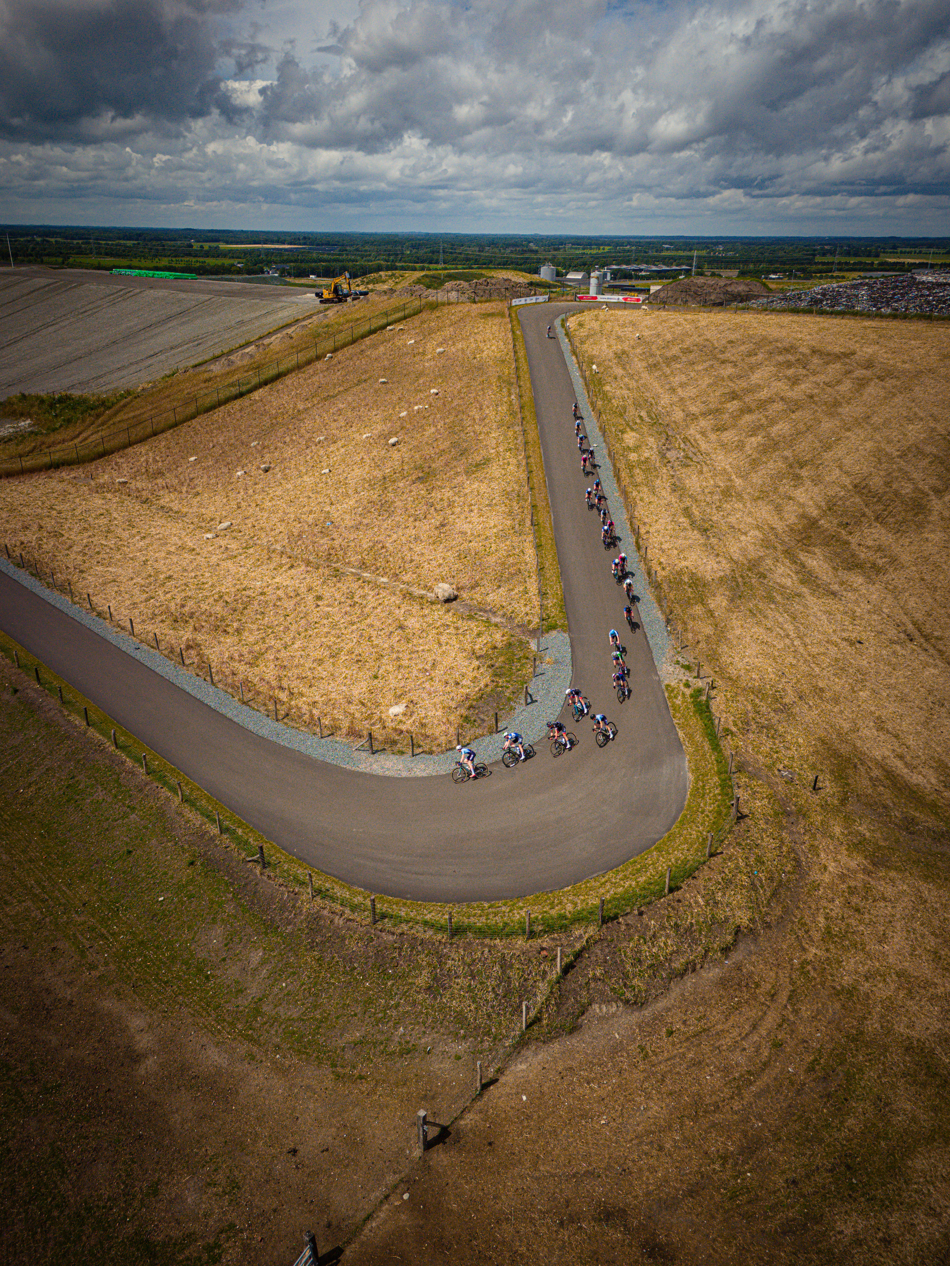 A group of cyclists riding a long, curved road. They are wearing numbers and helmets for safety. The sky is cloudy overhead.