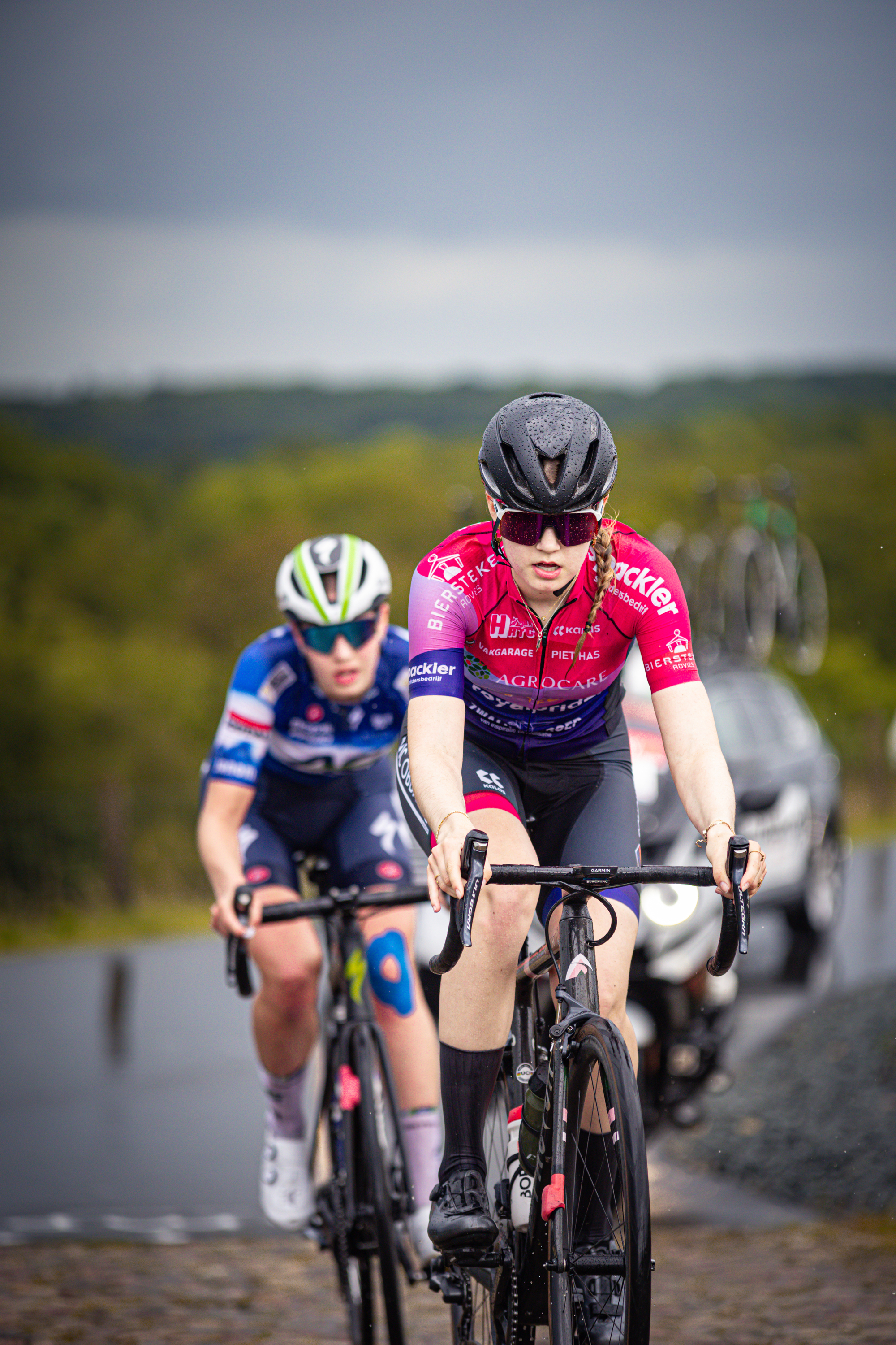 Two people are riding bikes. The person in front is wearing a pink jersey with "Nederlands Kampioenschap" printed on it.