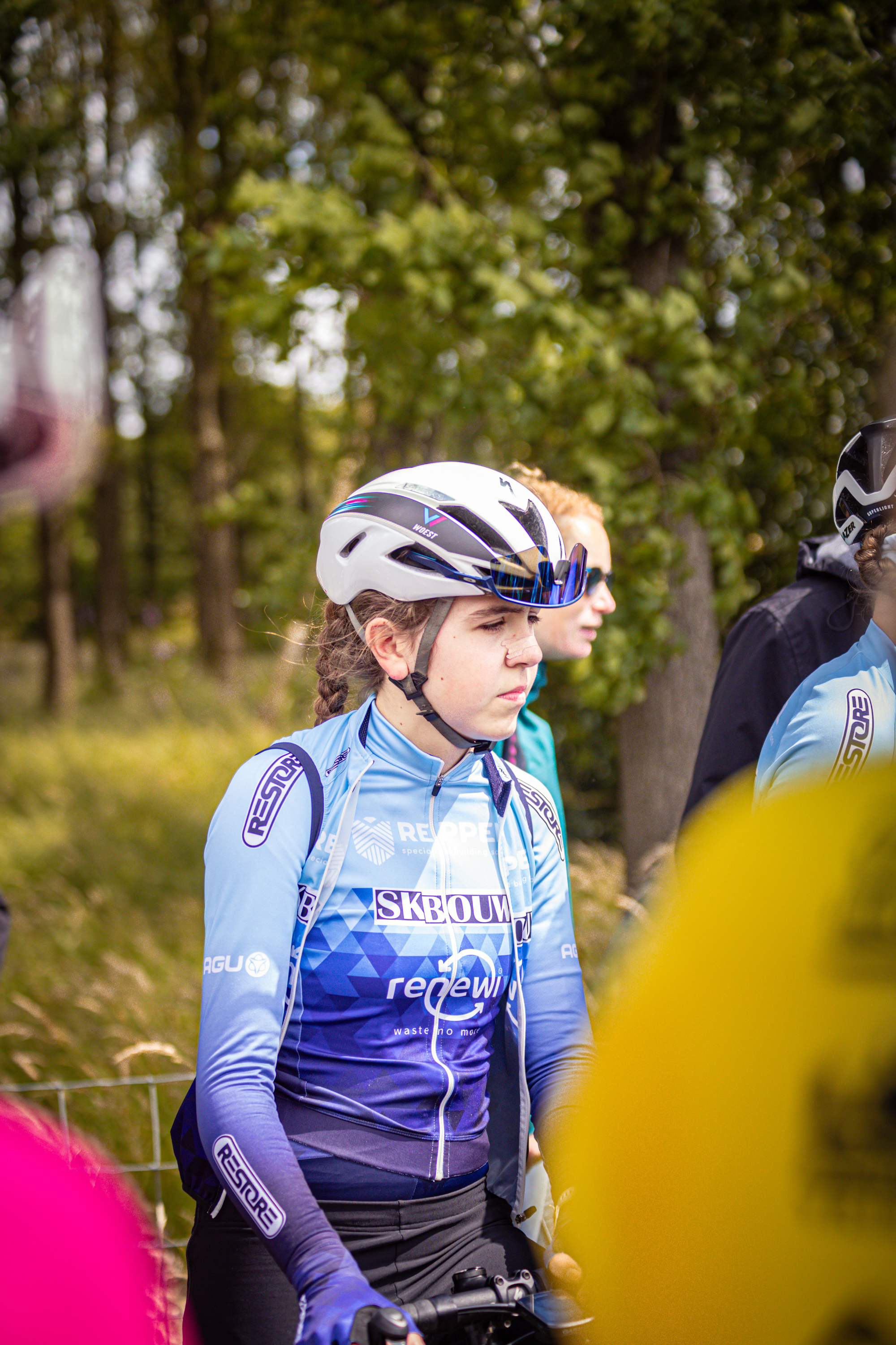 A young girl wearing a blue cycling jersey and a helmet stands on her bike, looking back.