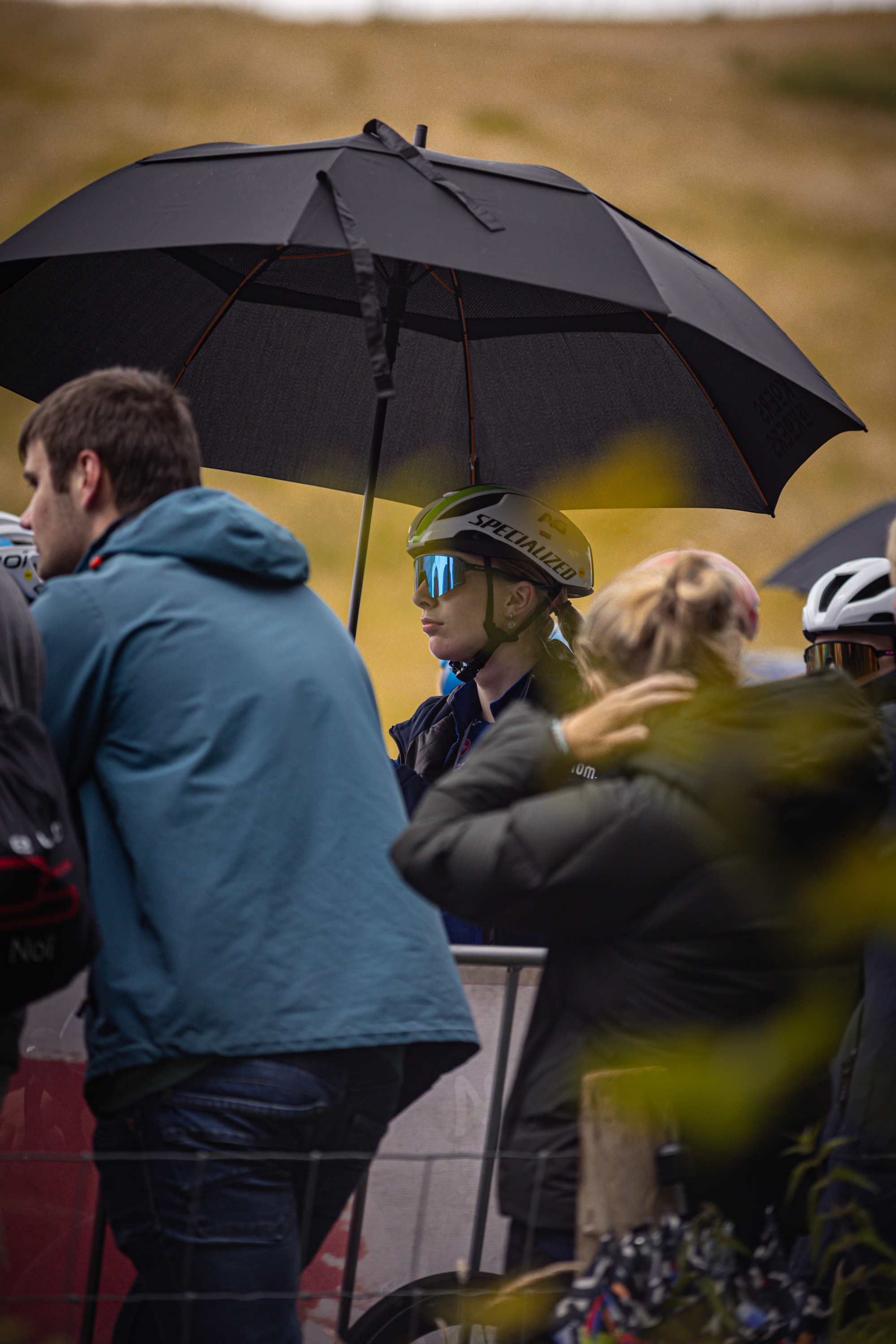 Two women wearing helmets and one man with a blue jacket stand next to each other under an umbrella.